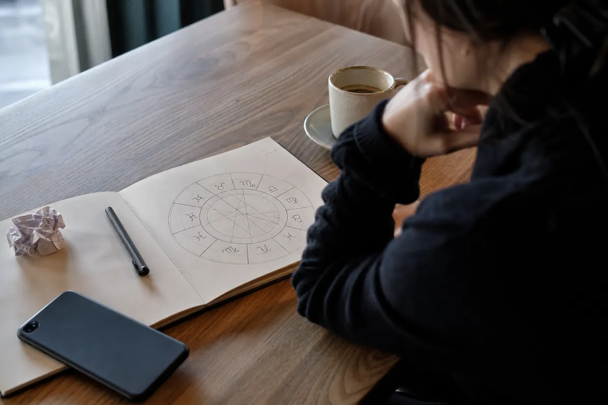 Persona pensierosa a un tavolo con taccuino aperto su un cerchio zodiacale, penna, telefono e tazza di caffè
