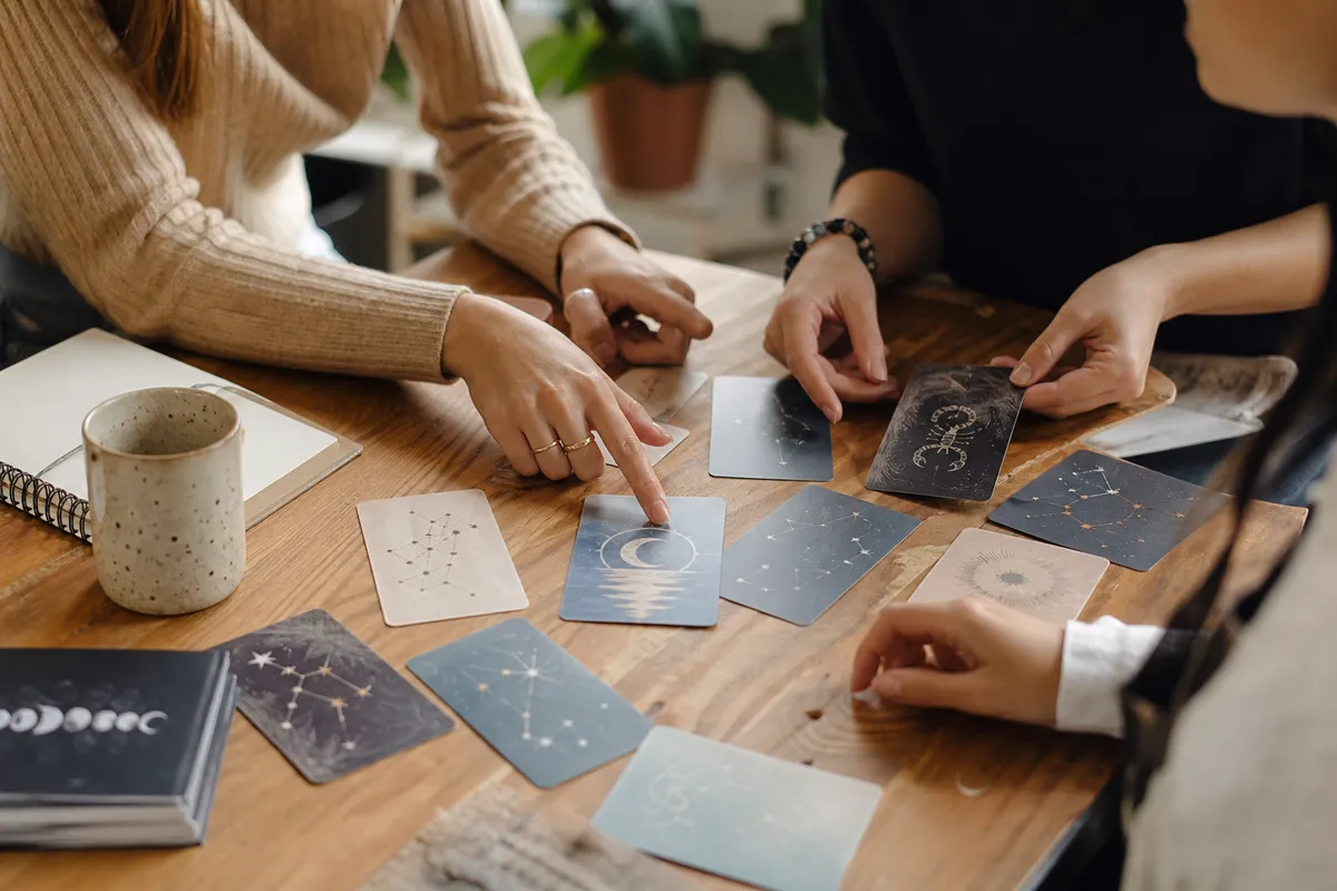 Mani attorno a un tavolo che dispongono carte con costellazioni e simboli lunari, accanto a tazza e quaderno
